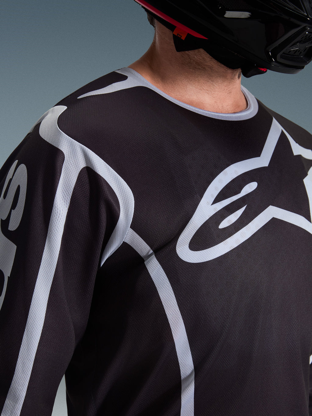 Alpinestars 2026 Fluid Apex Jersey, camisola de motocross, preta e cinzenta, com logótipo Alpinestars de grandes dimensões no peito, gola desportiva, usada com um capacete preto, equipamento de performance para condução todo-o-terreno