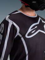 Alpinestars 2026 Fluid Apex Jersey, camisola de motocross, preta e cinzenta, com logótipo Alpinestars de grandes dimensões no peito, gola desportiva, usada com um capacete preto, equipamento de performance para condução todo-o-terreno