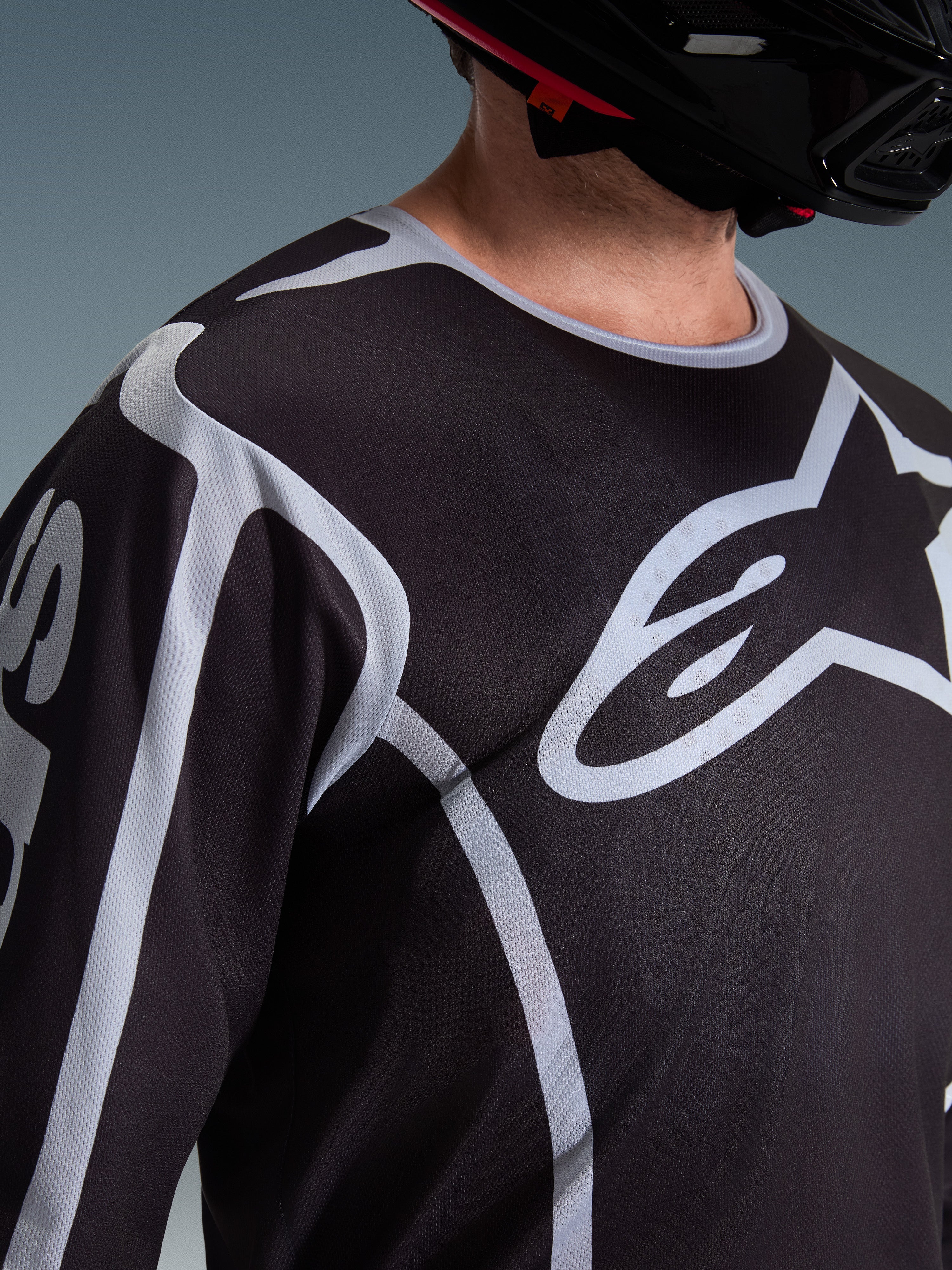 Alpinestars 2026 Fluid Apex Jersey, camisola de motocross, preta e cinzenta, com logótipo Alpinestars de grandes dimensões no peito, gola desportiva, usada com um capacete preto, equipamento de performance para condução todo-o-terreno