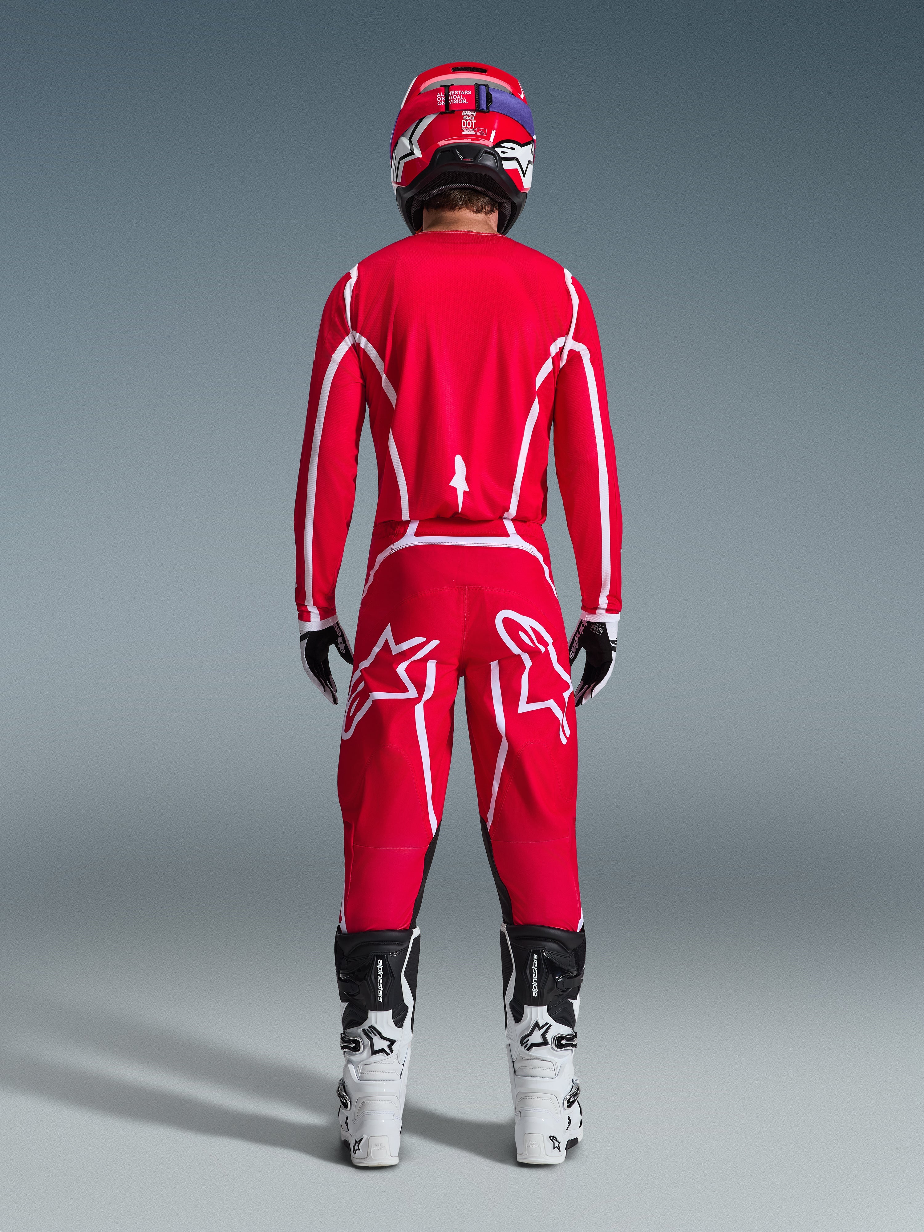 Rear view of a motocross rider wearing the Alpinestars 2026 Fluid Apex Gear Set, bright red jersey and pants with white stripes and logos, paired with white and black boots and a red helmet, professional off-road racing kit