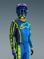 Piloto de motocross com a camisola Alpinestars 2026 Fluid Apex em azul e amarelo fluorescente, calças a condizer em azul e amarelo néon, capacete roxo e amarelo com óculos azuis e luvas coordenadas, equipamento completo de corrida todo-o-terreno