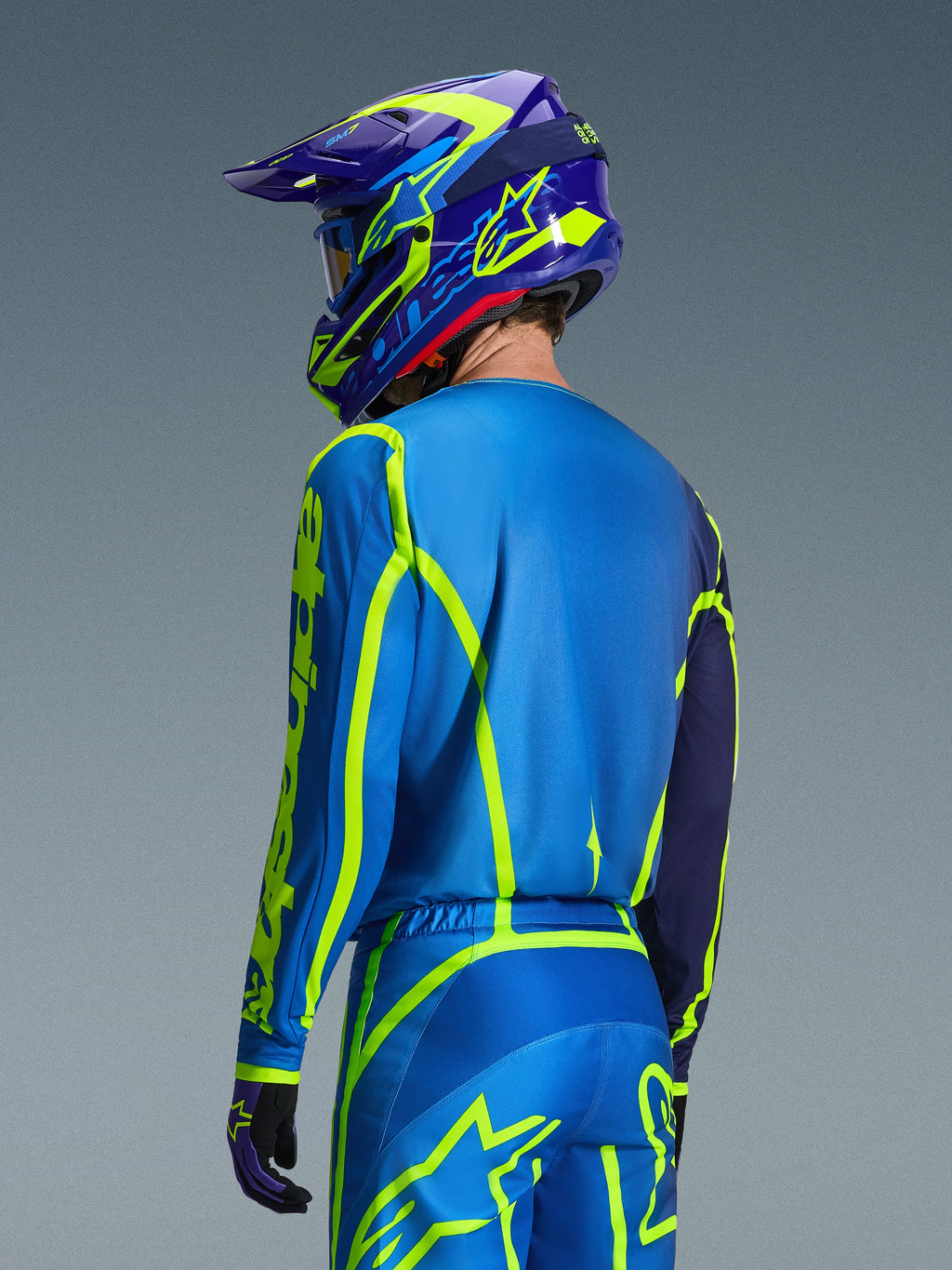 Alpinestars Camisola Fluid Apex 2026, Blue Yellow Fluo, vista traseira do piloto a usar calças de motocross e capacete a condizer, com gráficos geométricos em amarelo fluorescente e um ajuste atlético relaxado, equipamento de corrida off-road coordenado