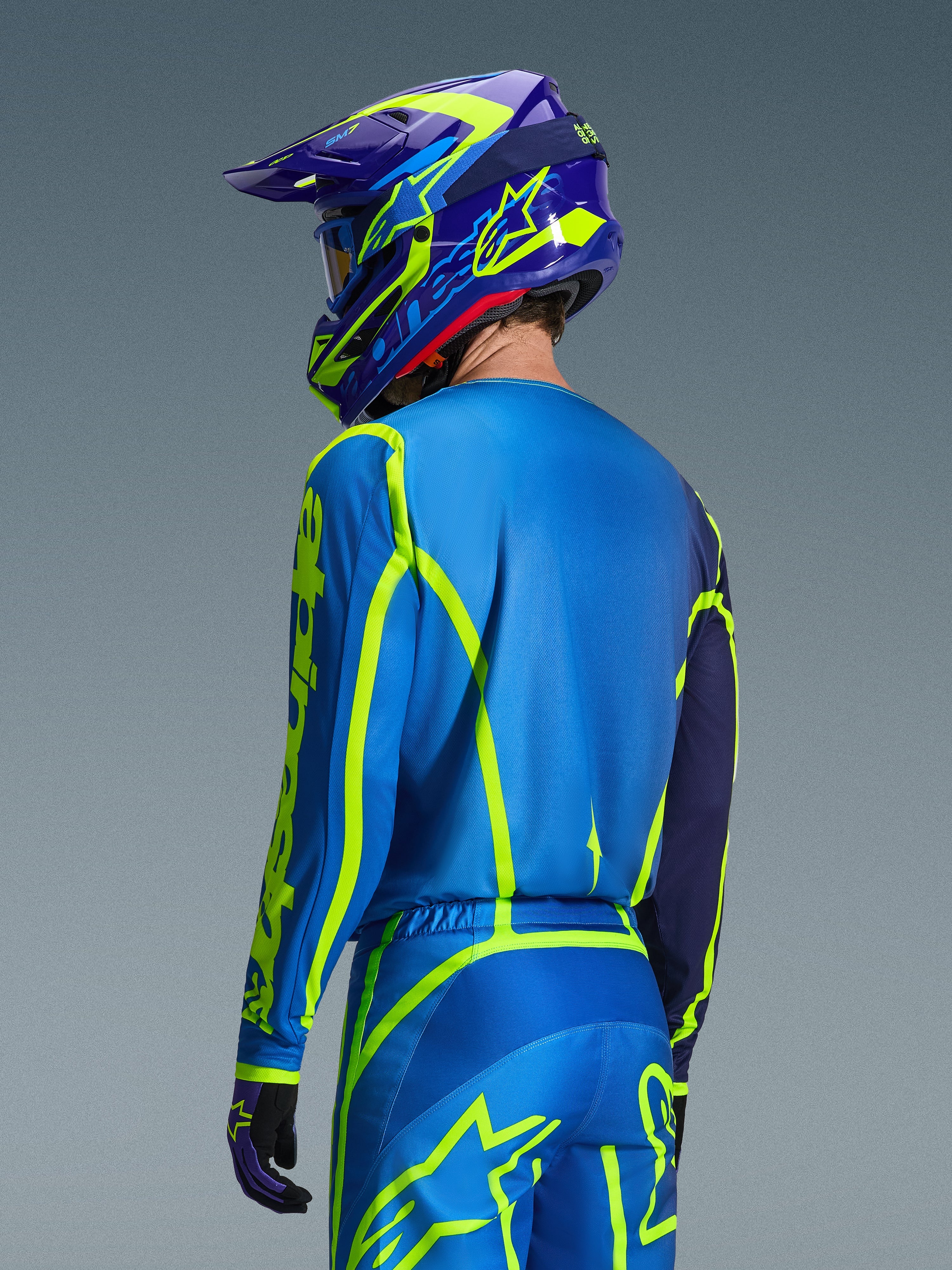 Alpinestars Camisola Fluid Apex 2026, Blue Yellow Fluo, vista traseira do piloto a usar calças de motocross e capacete a condizer, com gráficos geométricos em amarelo fluorescente e um ajuste atlético relaxado, equipamento de corrida off-road coordenado
