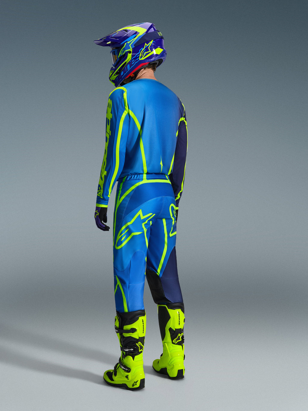 Piloto de motocross visto de costas a usar a Camisola Alpinestars 2026 Fluid Apex em Azul Amarelo Fluo, calças a condizer em azul e amarelo fluorescente, botas amarelo néon e capacete coordenado, equipamento profissional de corrida para todo-o-terreno