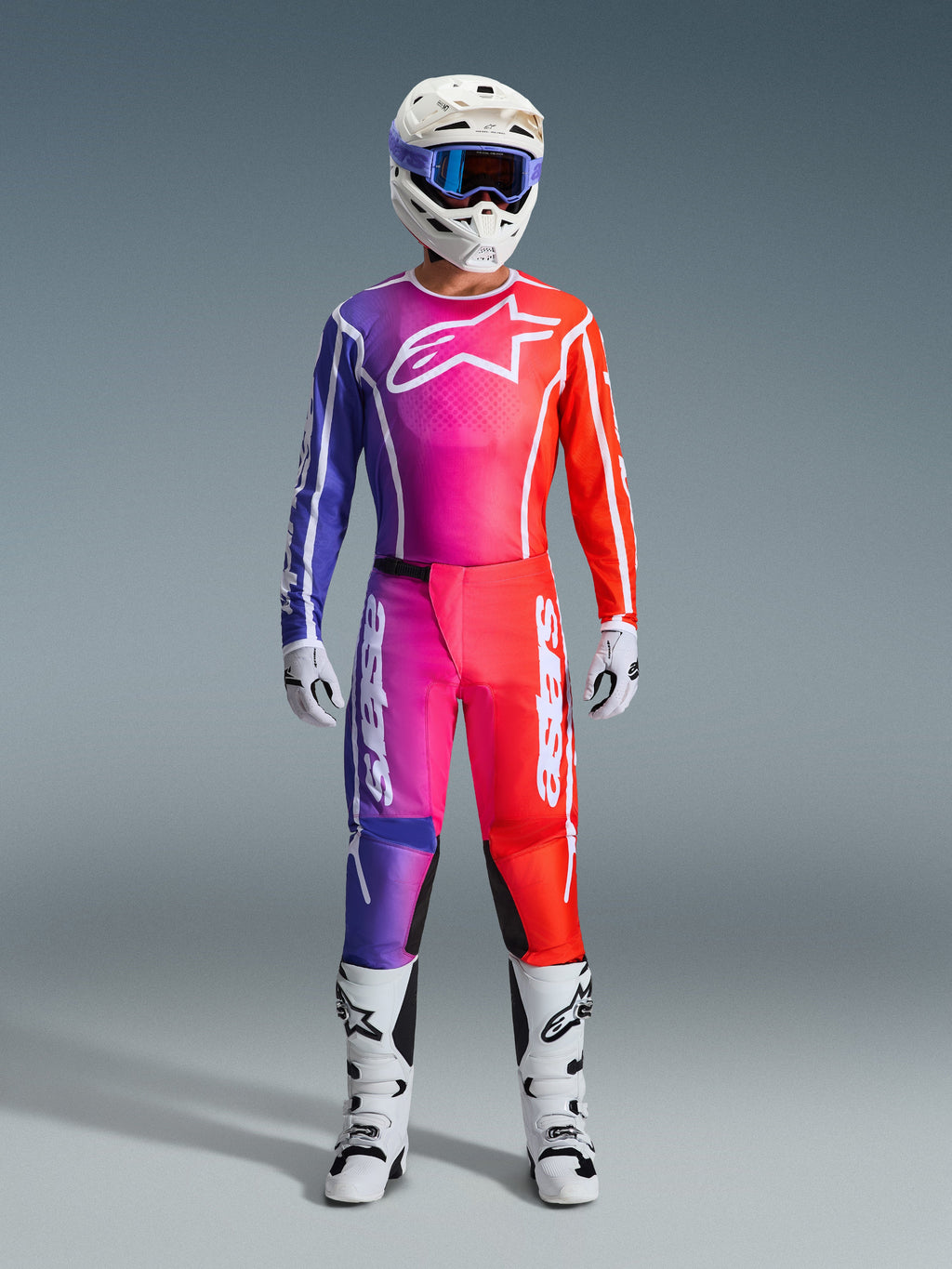 Equipamento completo de motocross Alpinestars 2026 Fluid Apex, camisola e calças com gradiente em roxo, rosa e laranja, logótipo Alpinestars branco no peito, capacete branco com óculos azuis e botas brancas, design para performance todo-o-terreno