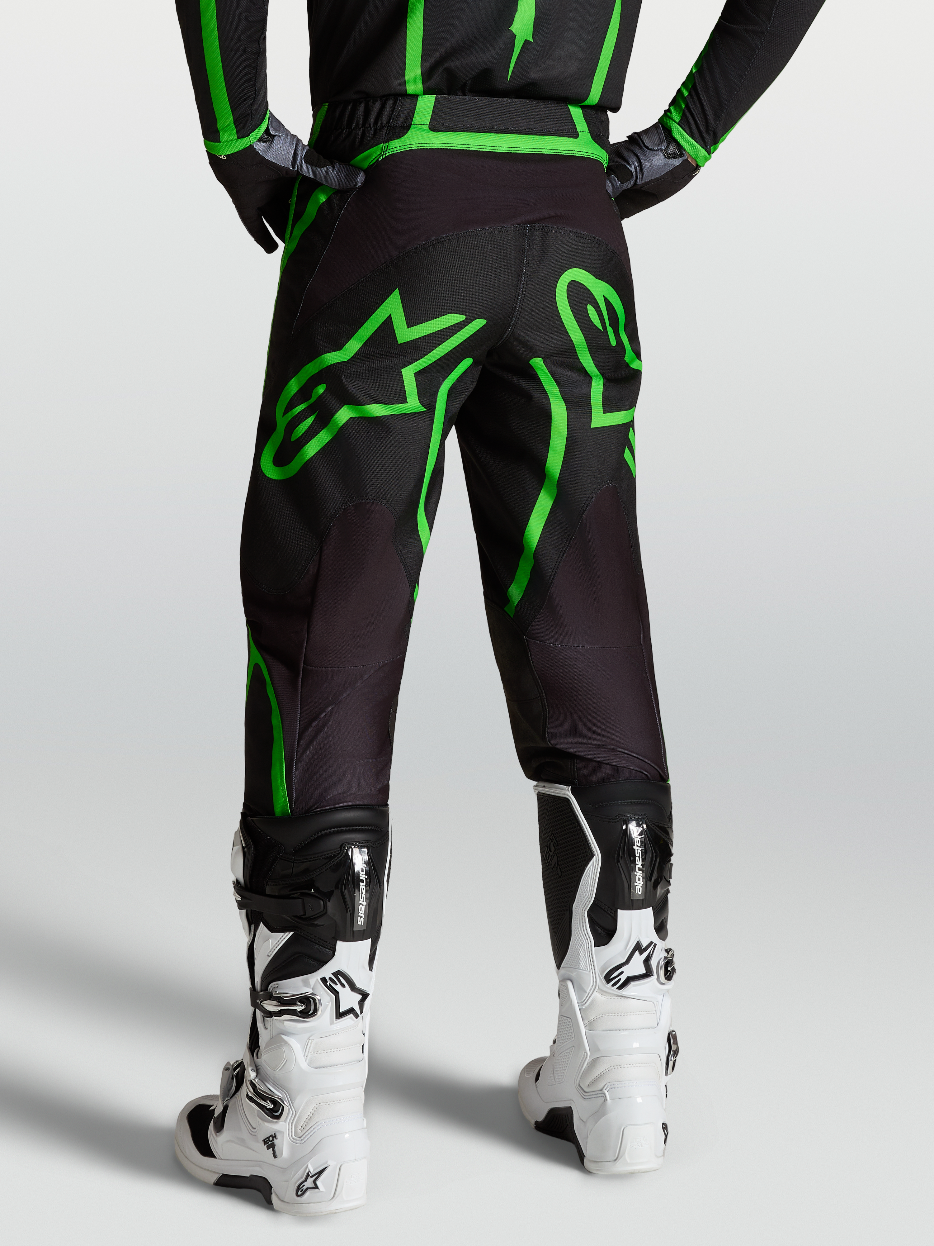 Alpinestars Fluid Apex V2, calças de motocross para homem, pretas e verdes fluo, vista traseira no modelo com botas brancas.