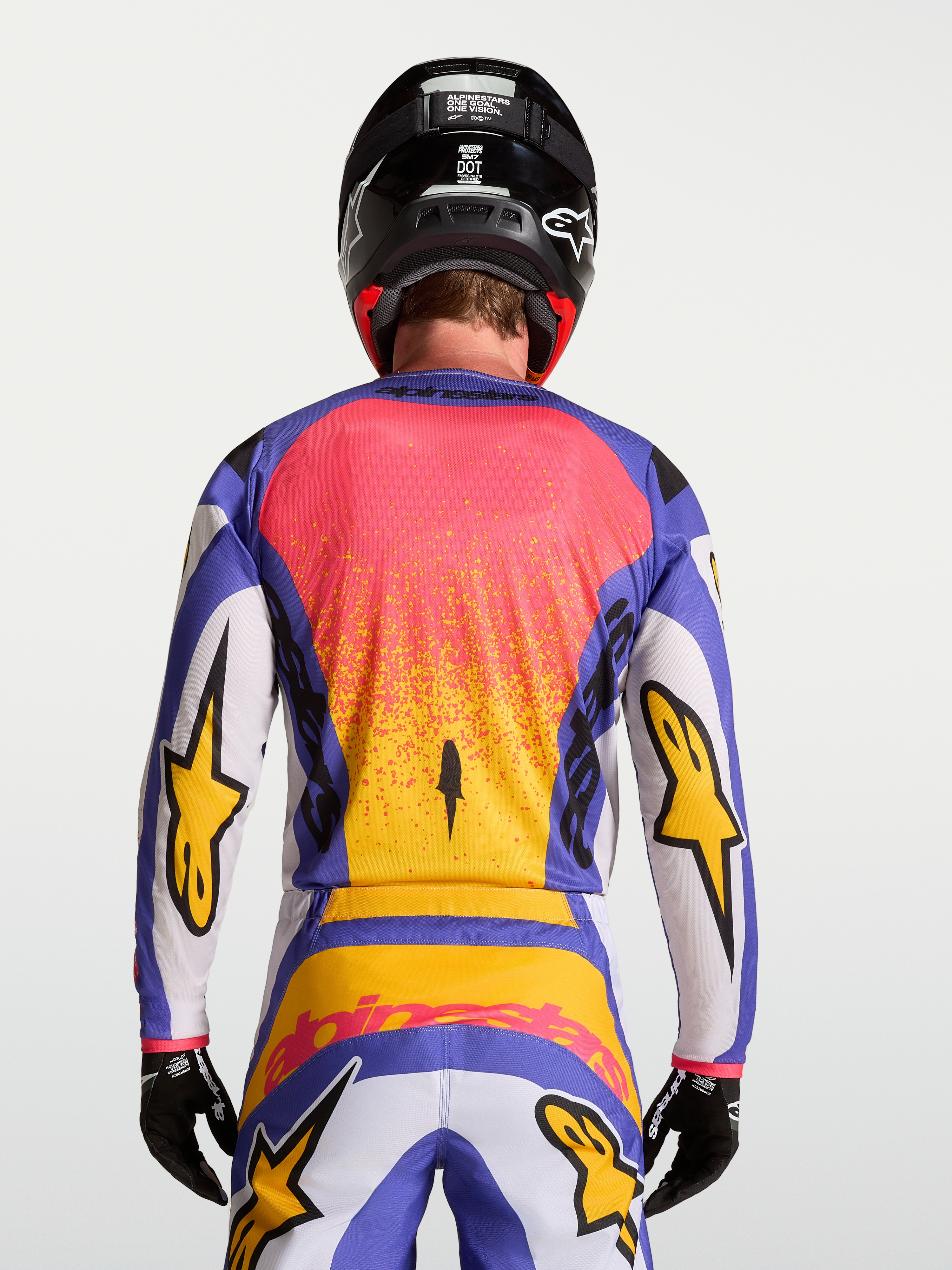 Alpinestars Camisola Fluid Drag, equipamento de motocross para homem em lilás e azul com detalhes dourados, vista traseira com calças e capacete a condizer.