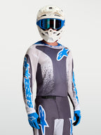 Camisola Alpinestars Fluid Drag, cinzenta e azul, equipamento de motocross para homem com capacete e luvas brancos.