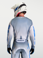 Alpinestars Camisola Racer Amber, camisola de motocross cinzenta e branca com pormenores em azul, vista traseira no modelo com capacete branco.