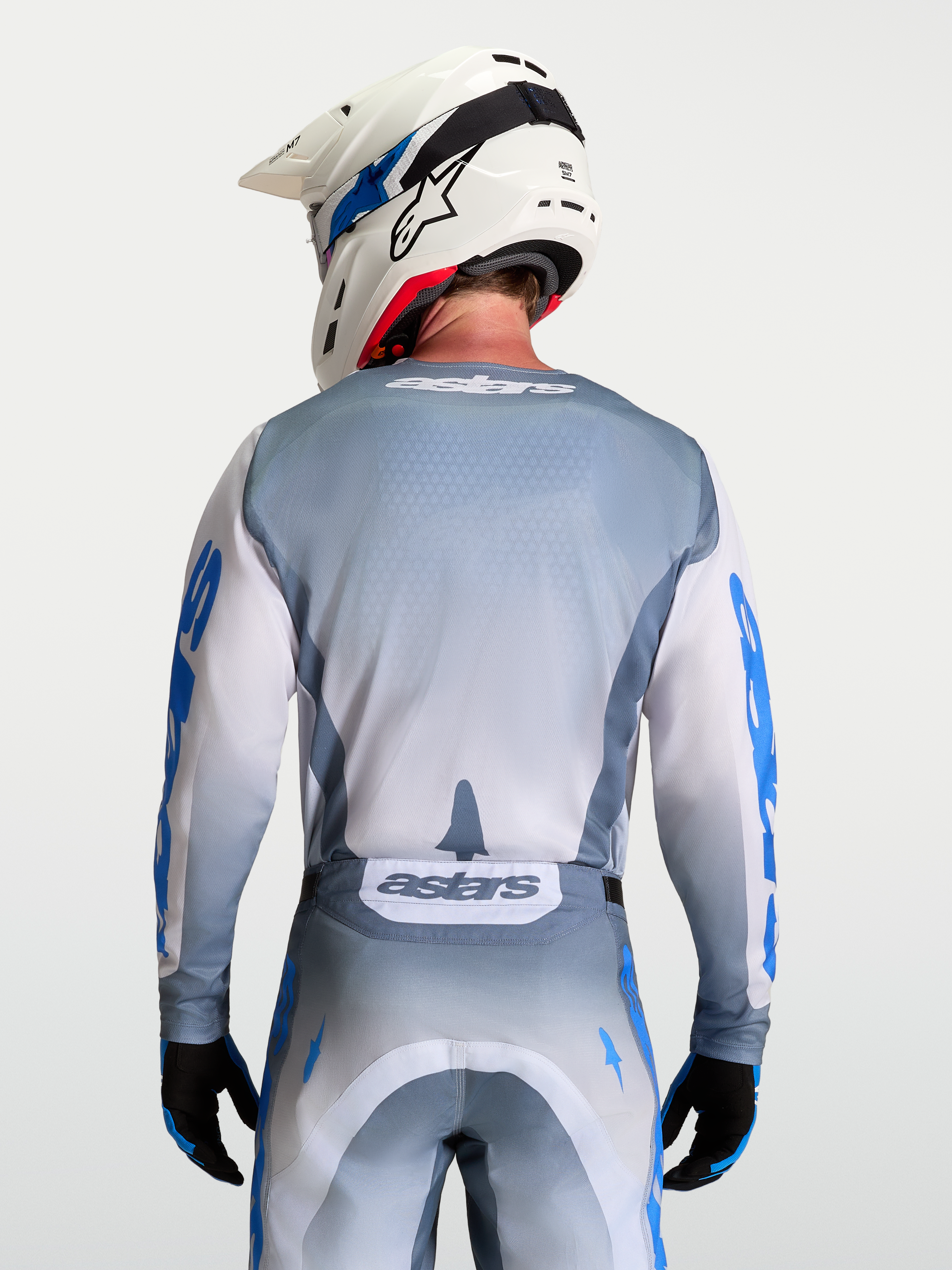 Alpinestars Camisola Racer Amber, camisola de motocross cinzenta e branca com pormenores em azul, vista traseira no modelo com capacete branco.