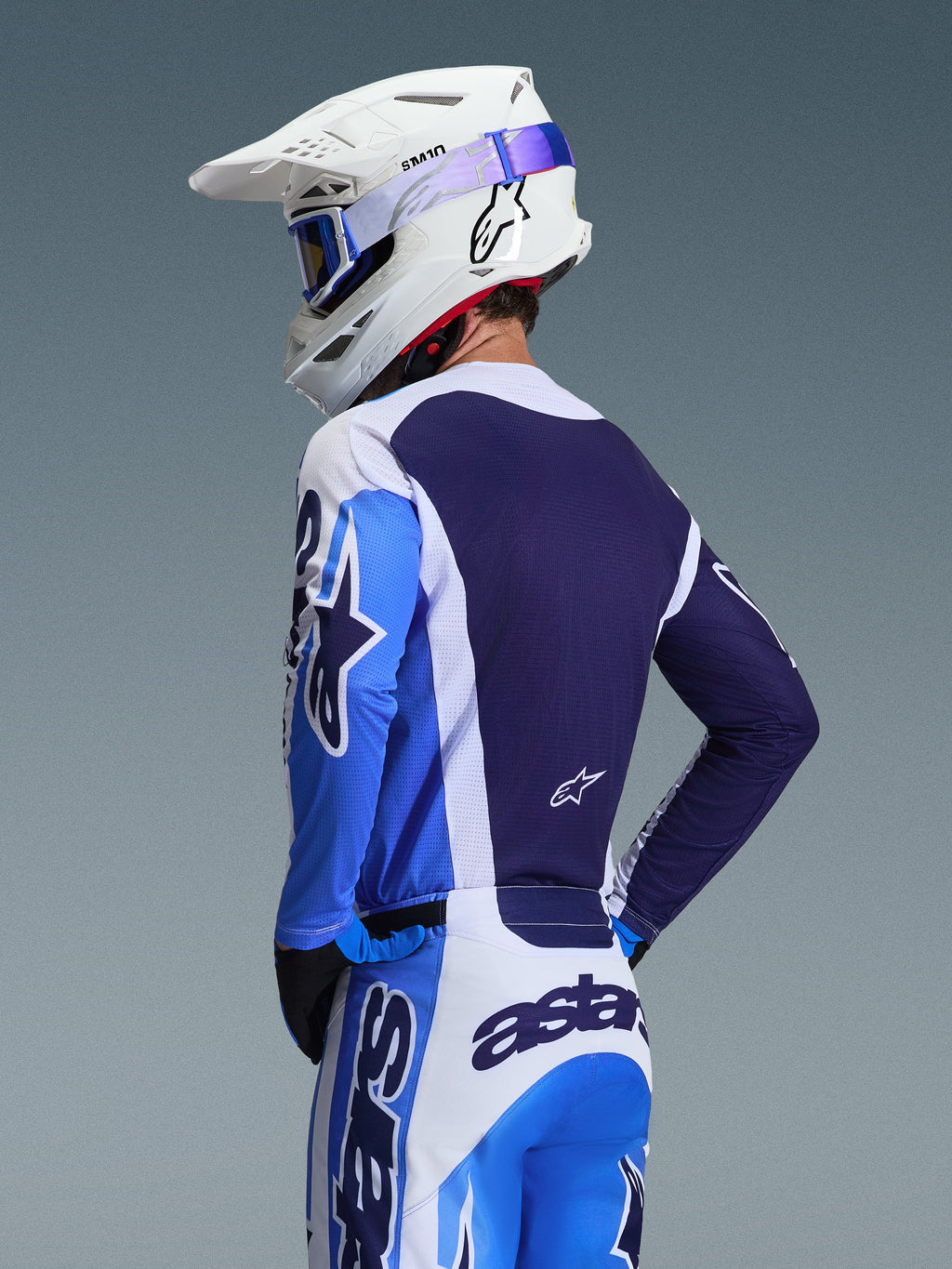 Piloto de motocross com camisola Alpinestars 2026 Racer Air Portl em azul Ucla e branco, vista traseira destacando o painel de malha respirável azul marinho, usada com calças a condizer e capacete Alpinestars branco com óculos azuis, conjunto de equipamento de motocross coordenado