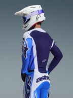Piloto de motocross com camisola Alpinestars 2026 Racer Air Portl em azul Ucla e branco, vista traseira destacando o painel de malha respirável azul marinho, usada com calças a condizer e capacete Alpinestars branco com óculos azuis, conjunto de equipamento de motocross coordenado