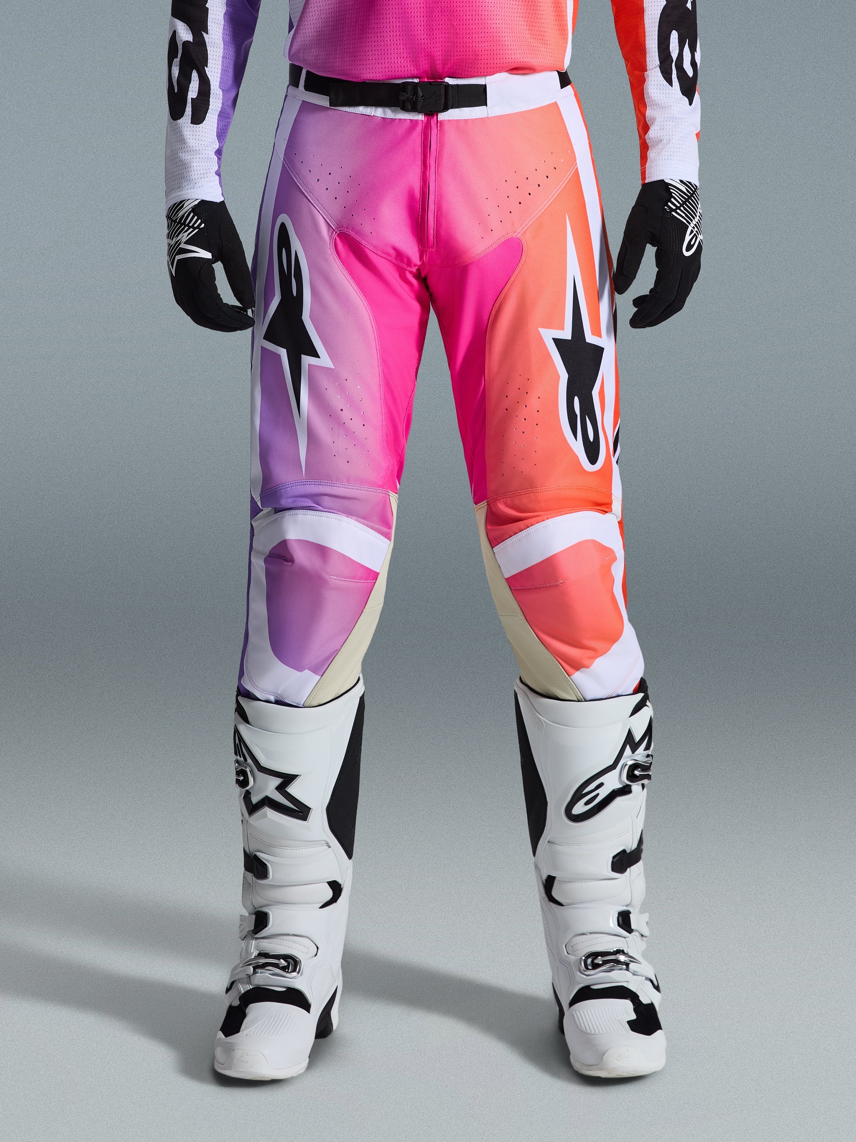 Alpinestars 2026 Racer Air Portl Pants, calças de motocross, gradiente vibrante de roxo, rosa e cor-de-laranja, com logótipos de estrela em destaque nas coxas, usadas com botas de corrida brancas e luvas pretas, concebidas para condução todo-o-terreno de alto desempenho.
