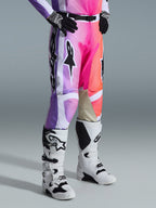 Alpinestars 2026 Racer Air Portl Pants, calças de motocross, grafismo multicolor com gradientes em roxo, cor-de-rosa e laranja, usadas com botas de motocross brancas e pretas e luvas pretas, com design de ajuste de corrida e painéis internos em couro bege nos joelhos
