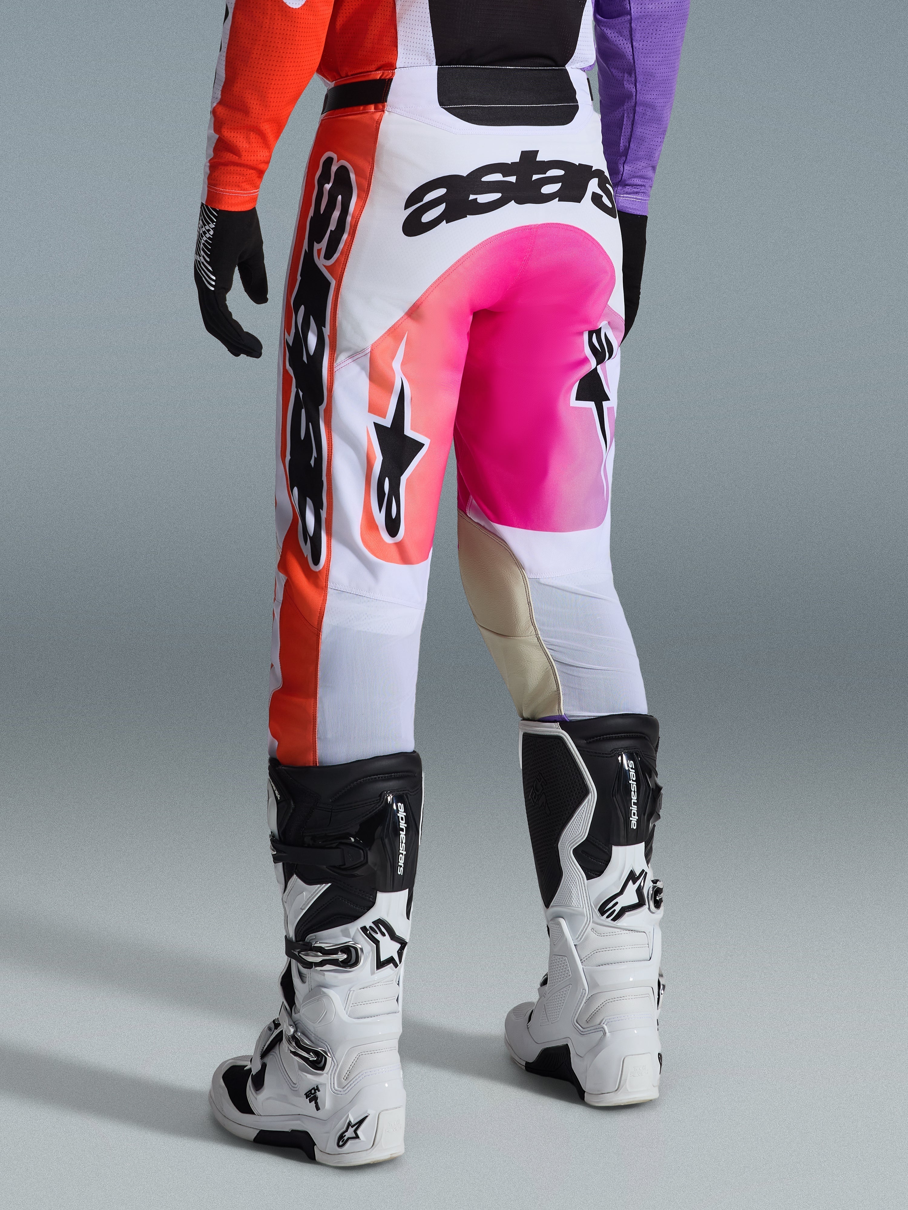 Alpinestars 2026 Racer Air Portl Pants, calças de motocross, multicolor com branco, cor-de-rosa vibrante e laranja, vista traseira com botas Alpinestars brancas e pretas, com logótipos marcantes e construção de competição durável