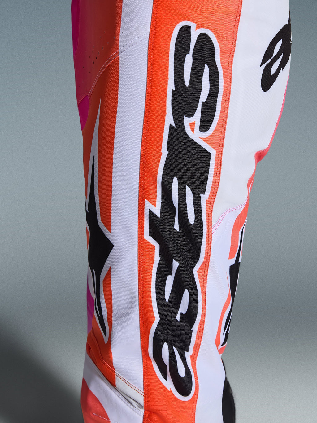 Alpinestars 2026 Racer Air Portl Pants, calças de motocross, cor-de-laranja, branco e rosa, grande plano mostrando a marca Alpinestars e o logótipo de estrela em preto, tecido perfurado para ventilação, vestuário de corrida off-road durável