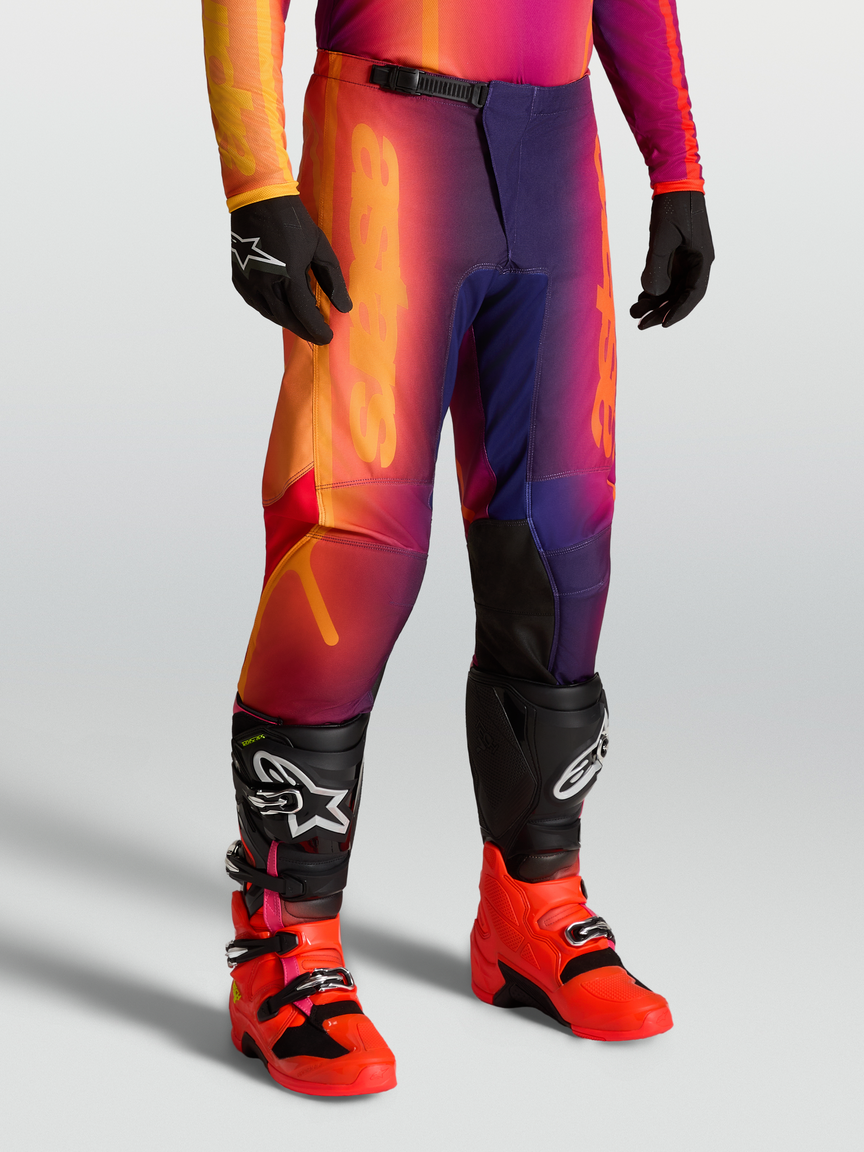 Alpinestars Calças Fluid Kepler, calças de motocross todo-o-terreno roxas e cor de laranja, ajuste desportivo com joelhos reforçados, usadas com botas e luvas pretas.