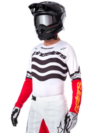 Motocross rider wearing the Alpinestars Limited Edition Techstar Deep Jersey, white jersey featuring black wavy stripes and red lower sleeves, styled with a black helmet, mirrored goggles, white and black gloves, and white racing pants, performance motocross outfit.