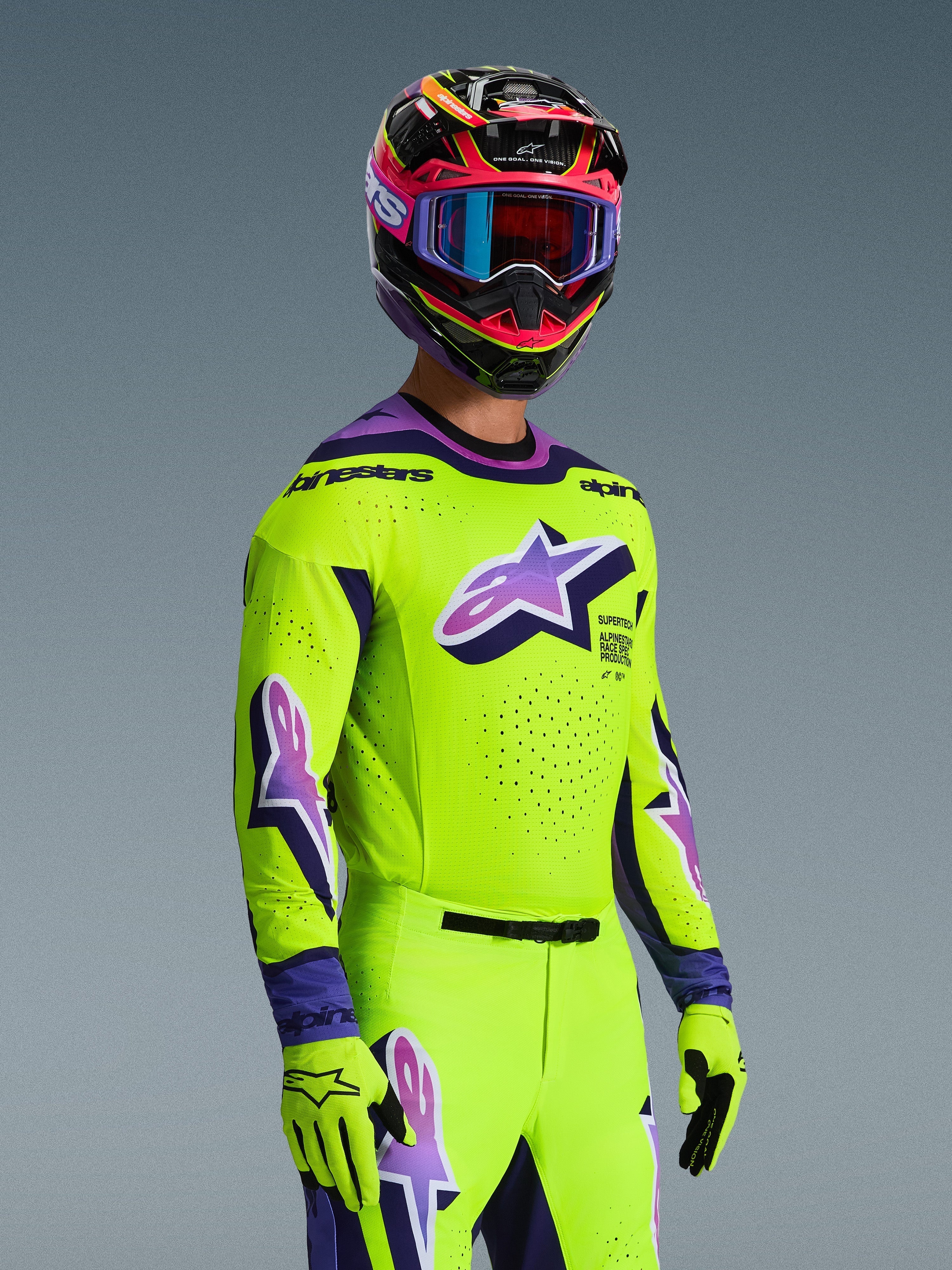 Motocross rider wearing the Alpinestars 2026 Supertech Vista Jersey and matching gear set, bright yellow with purple and black accents, featuring a large purple Astar logo on the chest and sleeves, matching gloves, and a multi-colored racing helmet with goggles, pro-level athletic fit for off-road racing.