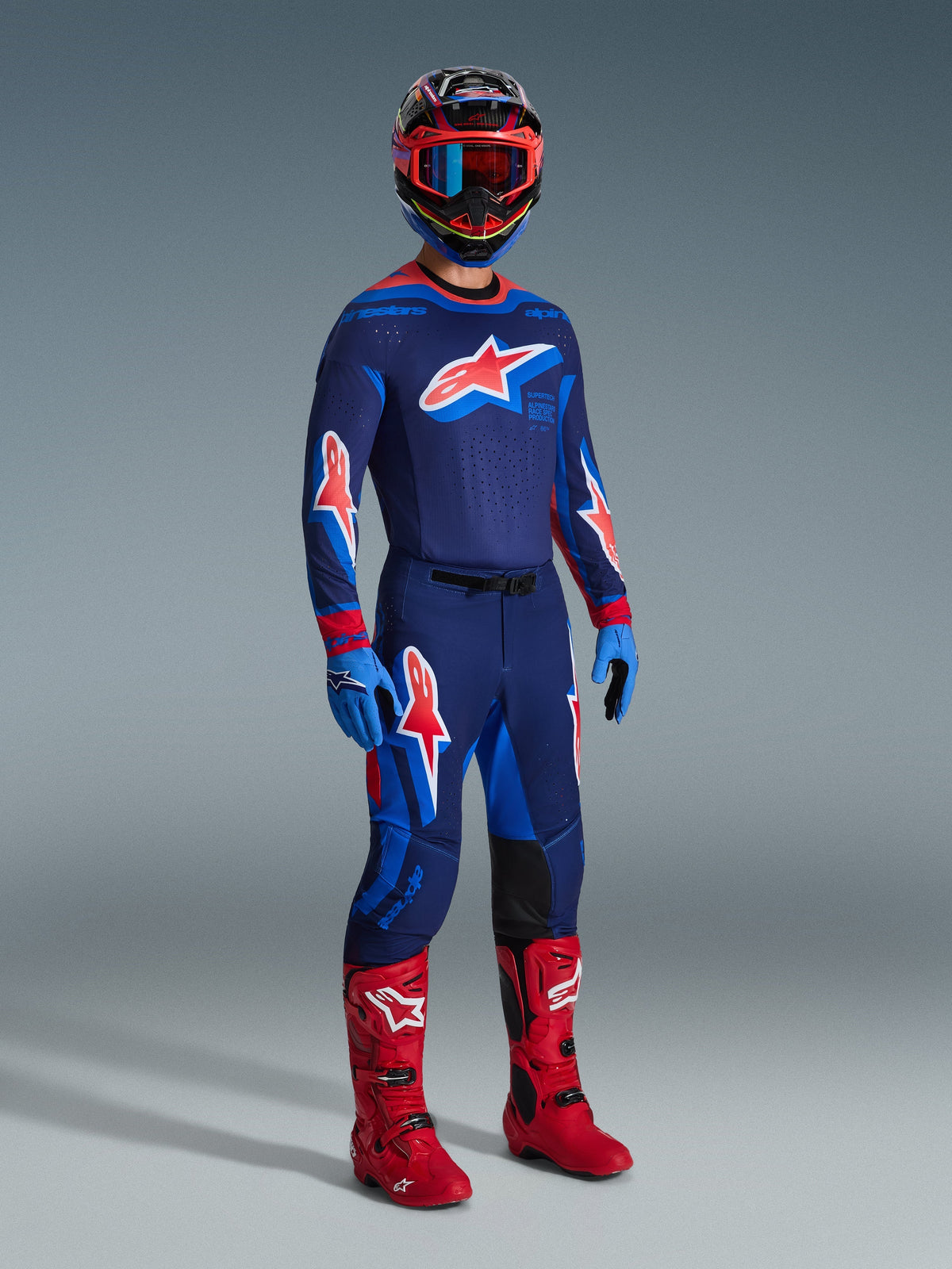 Piloto de motocross com equipamento de corrida completo, camisola Supertech Vista 2026 em azul-escuro com detalhes Hot Coral e Cobalt, calças de motocross a condizer, capacete multicolorido com óculos vermelhos, botas vermelho vivo, luvas azuis, conjunto coordenado para corridas todo-o-terreno