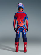 Motocross rider from rear view wearing Alpinestars 2026 Supertech Vista Gear Set in Dark Navy, Hot Coral, and Cobalt, featuring a vibrant red jersey back, bold branding on the pants, coordinated red boots, and a blue and pink helmet.