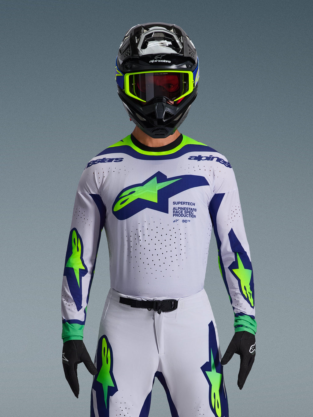 Conjunto de Equipamento Alpinestars 2026 Supertech Vista, camisola e calças de motocross, branco com grafismos em azul e verde néon, usado com capacete preto e óculos néon, com ventilação cortada a laser e ajuste de corrida atlético para competição profissional.
