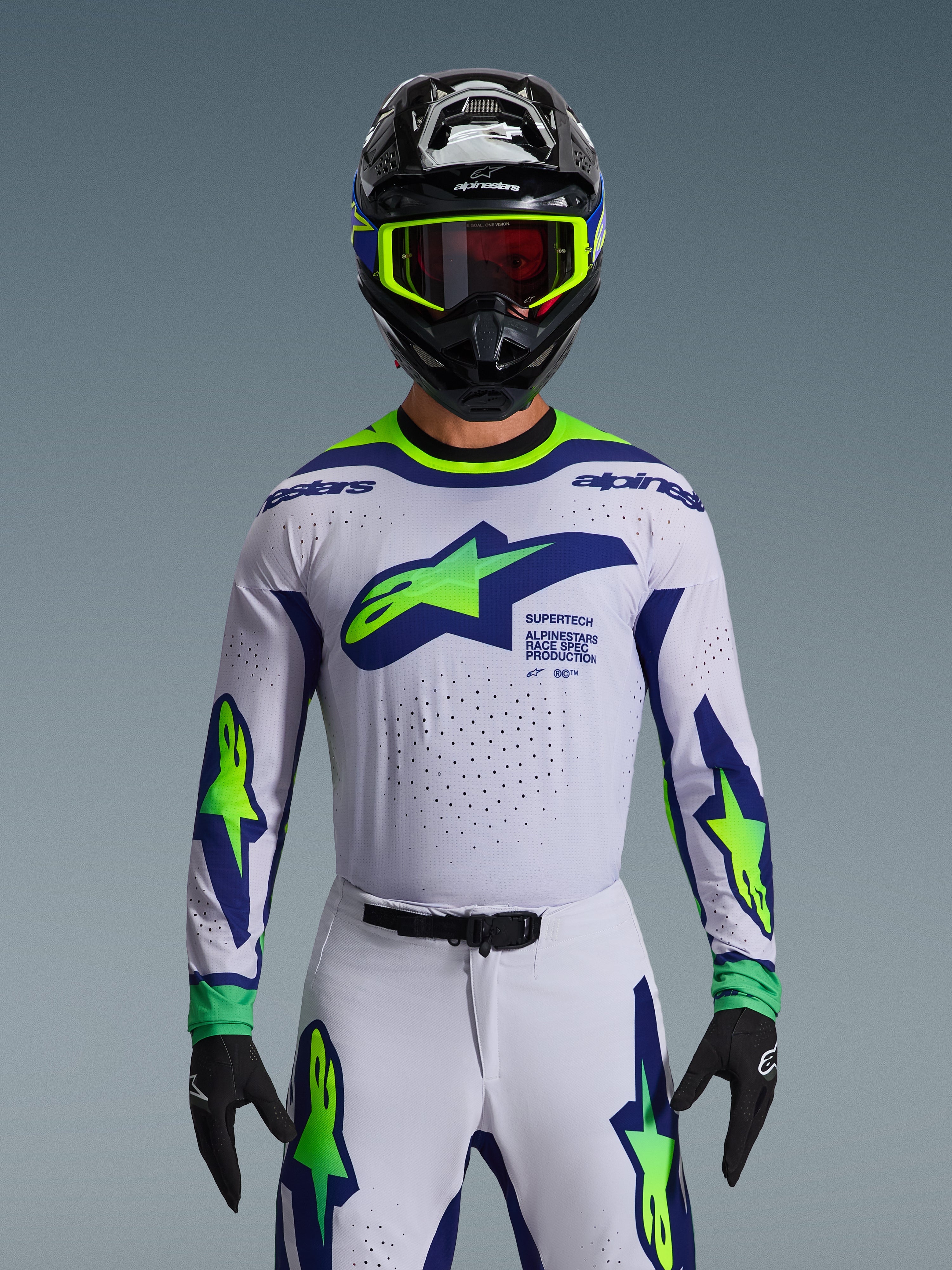 Conjunto de Equipamento Alpinestars 2026 Supertech Vista, camisola e calças de motocross, branco com grafismos em azul e verde néon, usado com capacete preto e óculos néon, com ventilação cortada a laser e ajuste de corrida atlético para competição profissional.