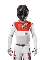 Piloto de motocross a usar a camisola Alpinestars 2024 Supertech Ward em White Hot Orange, camisola de malha elástica branca com um grafismo e logótipo vermelhos proeminentes no peito, capacete preto com óculos escuros, luvas brancas, equipamento de nível profissional para desempenho em todo-o-terreno