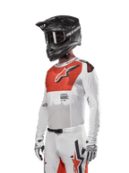 Camisola Alpinestars Supertech Ward 2024, camisola de motocross, branca e laranja vibrante, com painel peitoral vermelho e logótipo de estrela branco, mangas compridas com perfurações a laser, usada com capacete preto e luvas brancas, equipamento de motocross com ajuste atlético de competição