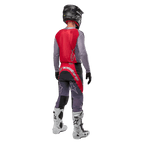 Alpinestars 2024 Supertech Dade Jersey, camisola de motocross, vermelho Iron Red Berry e cinzento, vista traseira num piloto, com painel traseiro vermelho, logótipo cinzento e mangas cinzentas, usada com calças de corrida coordenadas em cinzento e vermelho, botas brancas e capacete preto, equipamento profissional de todo-o-terreno.