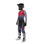 Piloto de motocross com equipamento de corrida completo, Camisola Alpinestars 2024 Supertech Dade em Iron Red Berry e cinzento, calças cinzentas com detalhes vermelhos, capacete preto com óculos, botas brancas e pretas, luvas pretas, conjunto coordenado para corridas todo-o-terreno