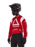 Alpinestars Techdura Jersey, camisola de motocross, vermelha e branca, com um design de triângulo branco e branding ASTARS, usada com capacete e luvas pretos, equipamento de corrida todo-o-terreno durável e com absorção de humidade