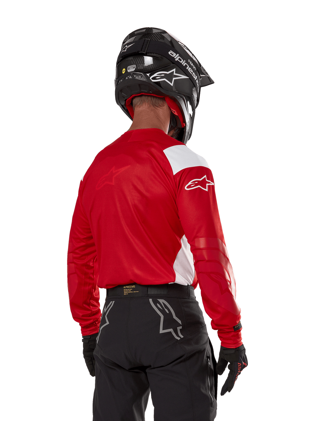 Piloto de motocross visto de costas a usar a camisola Alpinestars Techdura em vermelho vivo com painéis brancos, combinada com calças de todo-o-terreno pretas, capacete preto e luvas pretas, equipamento coordenado para a prática de enduro e motocross.