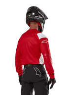 Piloto de motocross visto de costas a usar a camisola Alpinestars Techdura em vermelho vivo com painéis brancos, combinada com calças de todo-o-terreno pretas, capacete preto e luvas pretas, equipamento coordenado para a prática de enduro e motocross.