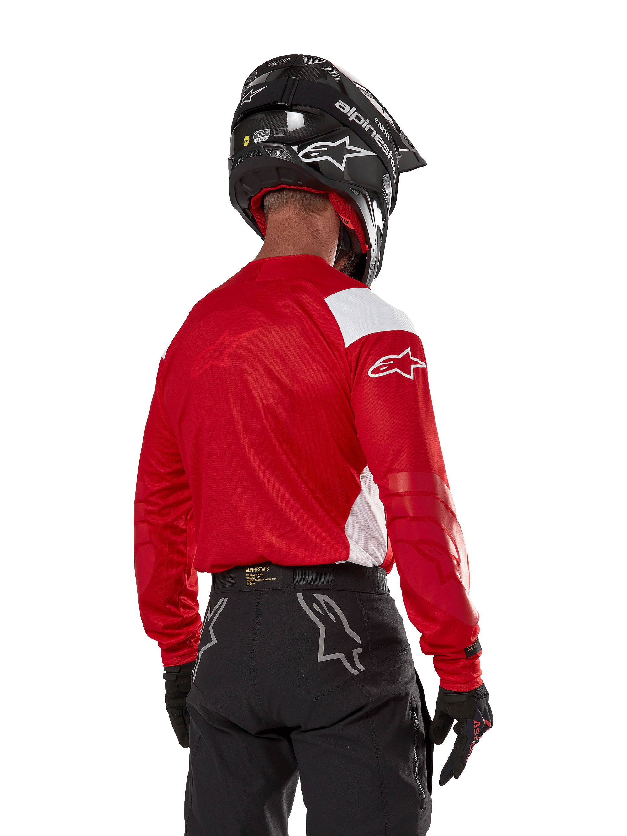Piloto de motocross visto de costas a usar a camisola Alpinestars Techdura em vermelho vivo com painéis brancos, combinada com calças de todo-o-terreno pretas, capacete preto e luvas pretas, equipamento coordenado para a prática de enduro e motocross.