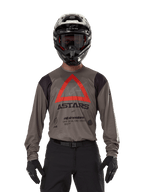 Piloto de motocross a usar a Camisola Alpinestars Techdura em Falcon Brown, com um triângulo vermelho proeminente e a marca 'ASTARS' no peito, acompanhada de calças pretas, capacete preto com óculos e luvas pretas, equipamento completo de corrida enduro