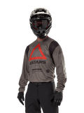 Alpinestars Techdura Jersey, camisola de motocross e todo-o-terreno, castanho Falcon com triângulo vermelho e logótipos pretos, usada com calças pretas, capacete preto e branco e luvas pretas