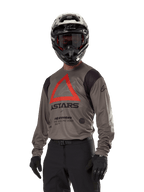 Alpinestars Techdura Jersey, camisola de motocross e todo-o-terreno, castanho Falcon com triângulo vermelho e logótipos pretos, usada com calças pretas, capacete preto e branco e luvas pretas