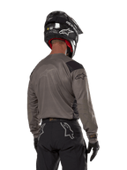 Piloto de motocross visto de costas a usar a camisola Alpinestars Techdura em Falcon Brown, combinada com calças de off-road pretas e um capacete preto, com um design gráfico moderno e parte traseira alongada para a prática de enduro