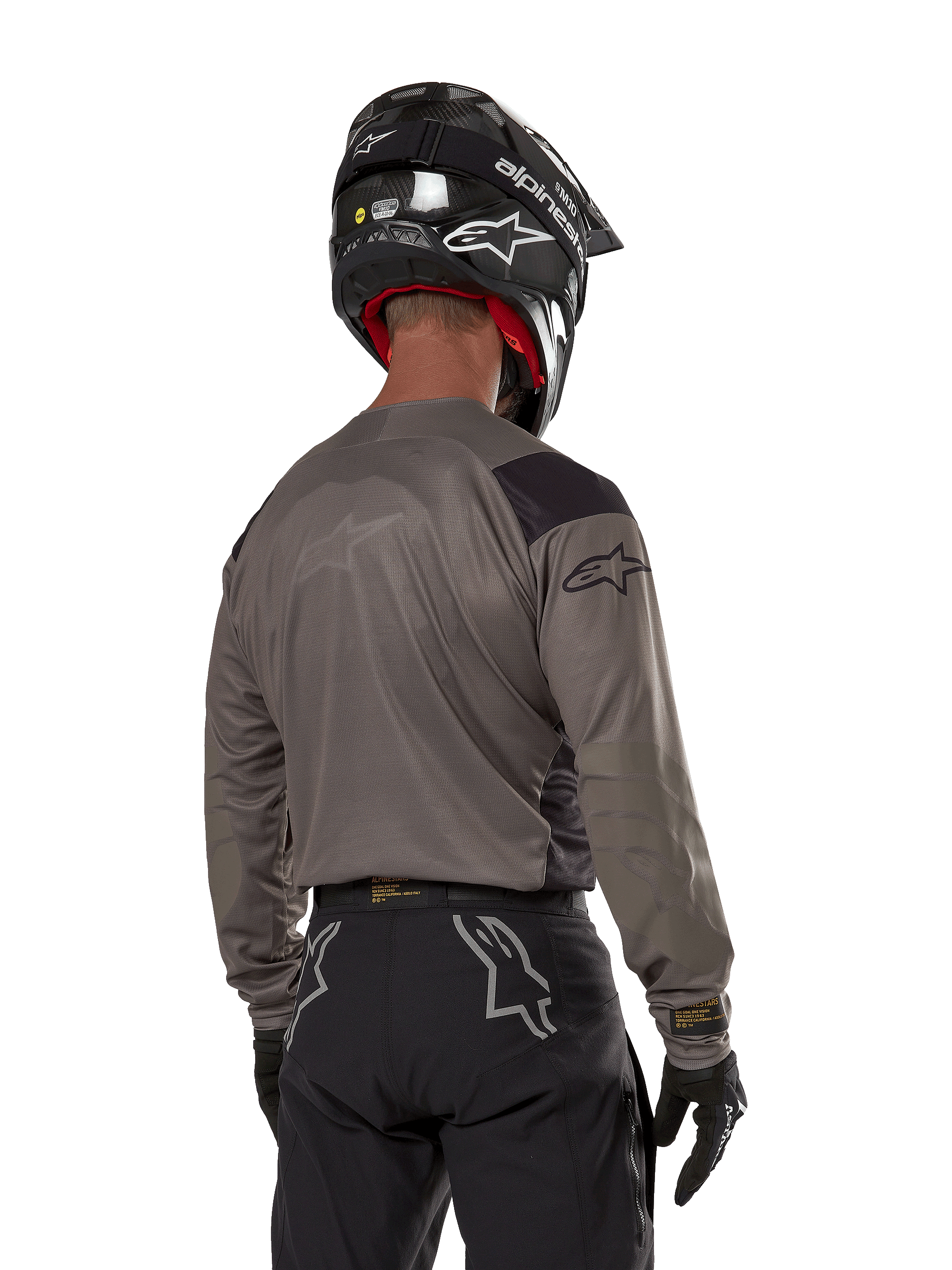Piloto de motocross visto de costas a usar a camisola Alpinestars Techdura em Falcon Brown, combinada com calças de off-road pretas e um capacete preto, com um design gráfico moderno e parte traseira alongada para a prática de enduro