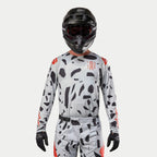 Alpinestars 2024 Techstar Rantera Jersey, camisola de motocross, camuflado cinzento com padrões pretos irregulares e detalhes vermelhos, usada por um piloto com calças a condizer, capacete preto e luvas, equipamento de corrida todo-o-terreno