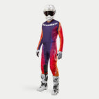 Equipamento completo de motocross Alpinestars, incluindo a camisola e calças 2024 Techstar Pneuma em roxo escuro, laranja e azul profundo, capacete branco, óculos de proteção, luvas brancas e pretas, e botas brancas com detalhes pretos, kit de alta performance.