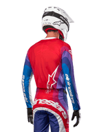 Piloto de motocross visto de costas a usar a camisola Alpinestars 2024 Techstar Pneuma em azul, vermelho e branco, com as costas vermelhas e logótipo de estrela branco, mangas azuis e roxas com a marca a branco, coordenada com calças a condizer e um capacete branco para corridas todo-o-terreno.