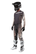 Piloto de motocross com equipamento completo de corrida, camisola Techstar Pneuma 2024 em Dark Sand e Iron Dust Gray, calças de motocross com padrão a condizer, capacete preto com óculos, botas brancas e pretas, luvas pretas, conjunto coordenado para competição off-road