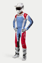 Piloto de motocross com equipamento de corrida completo, incluindo a Camisola Alpinestars 2024 Techstar Ocuri em azul claro, vermelho Mars e branco, calças a condizer, capacete branco com óculos, botas brancas e luvas brancas e pretas.