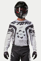 Piloto de motocross a vestir a camisola Alpinestars Racer Hana 2024 em Branco e Preto, combinada com capacete preto e calças de motocross pretas, com grafismos 'ASTARS' pretos arrojados e um logótipo central circular, equipamento profissional para todo-o-terreno