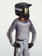 Alpinestars camisola Fluid Amber Jovem 2027, camisola de motocross em cinzento e areia com capacete de corrida preto.