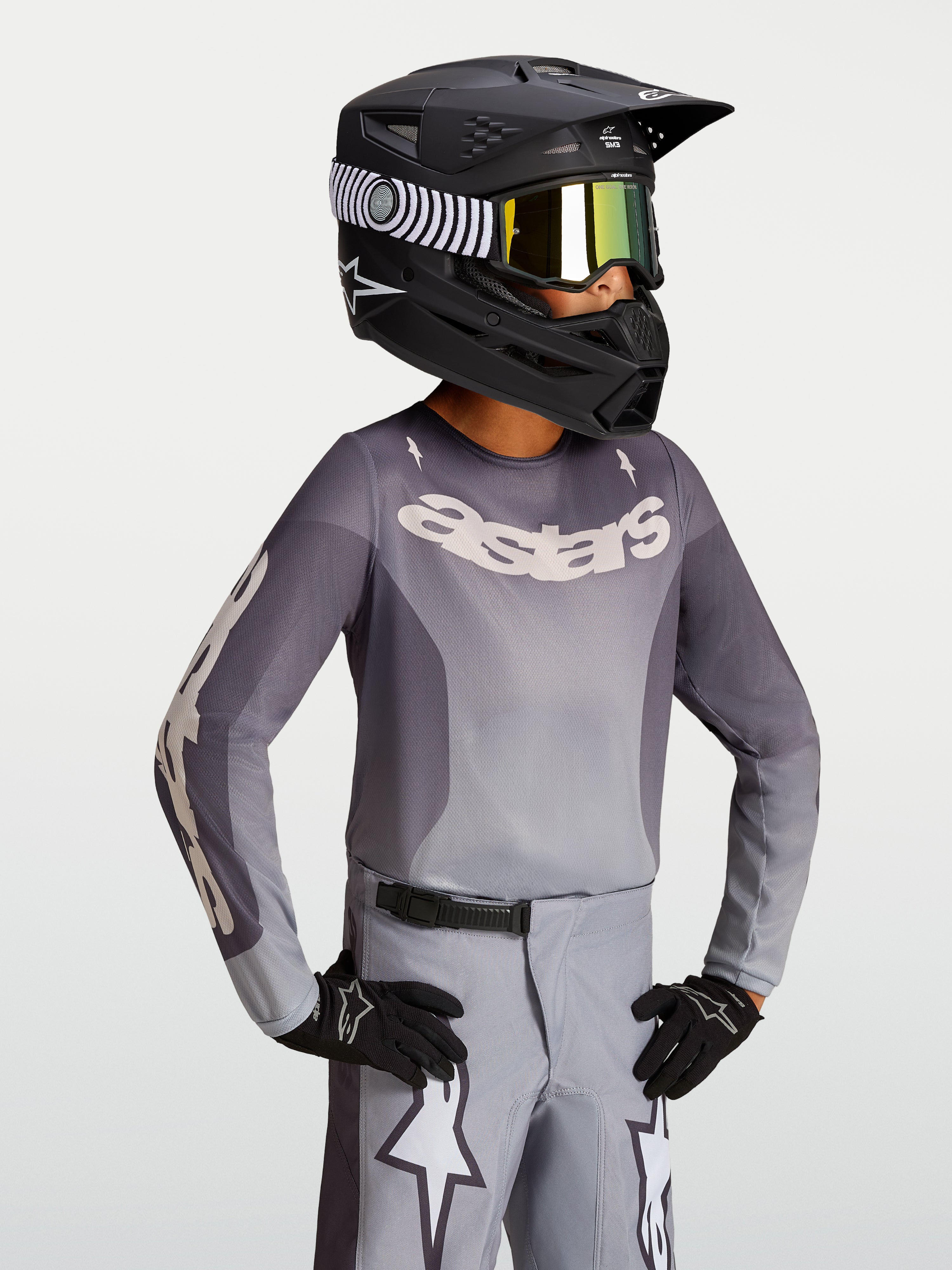 Alpinestars camisola Fluid Amber Jovem 2027, camisola de motocross em cinzento e areia com capacete de corrida preto.