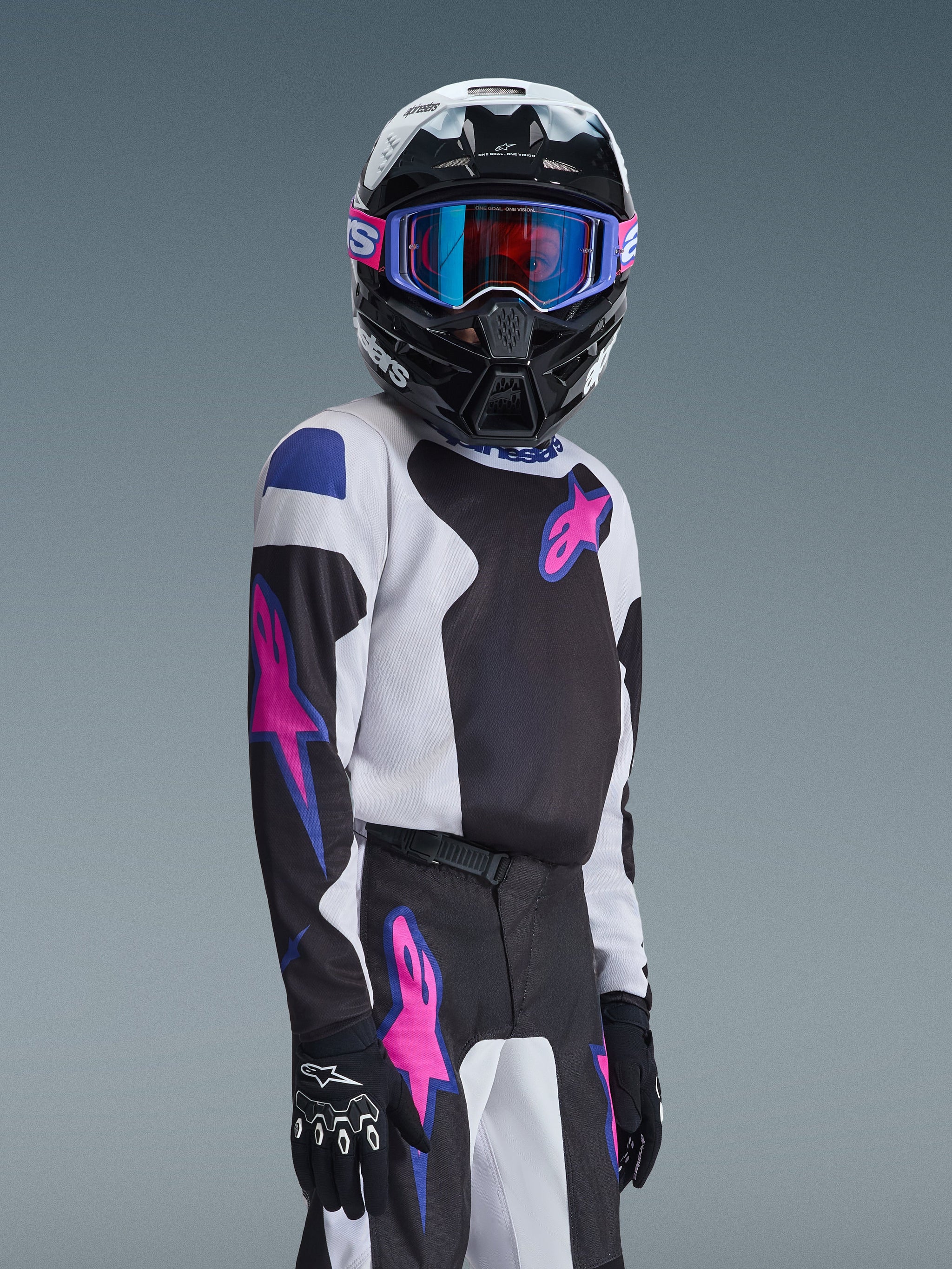 2026 Youth Fluid Grid Jersey worn by a young motocross rider, black and white jersey with vibrant purple and pink Alpinestars logos, matching pants, black and white helmet with iridescent goggles, professional off-road racing kit for youth