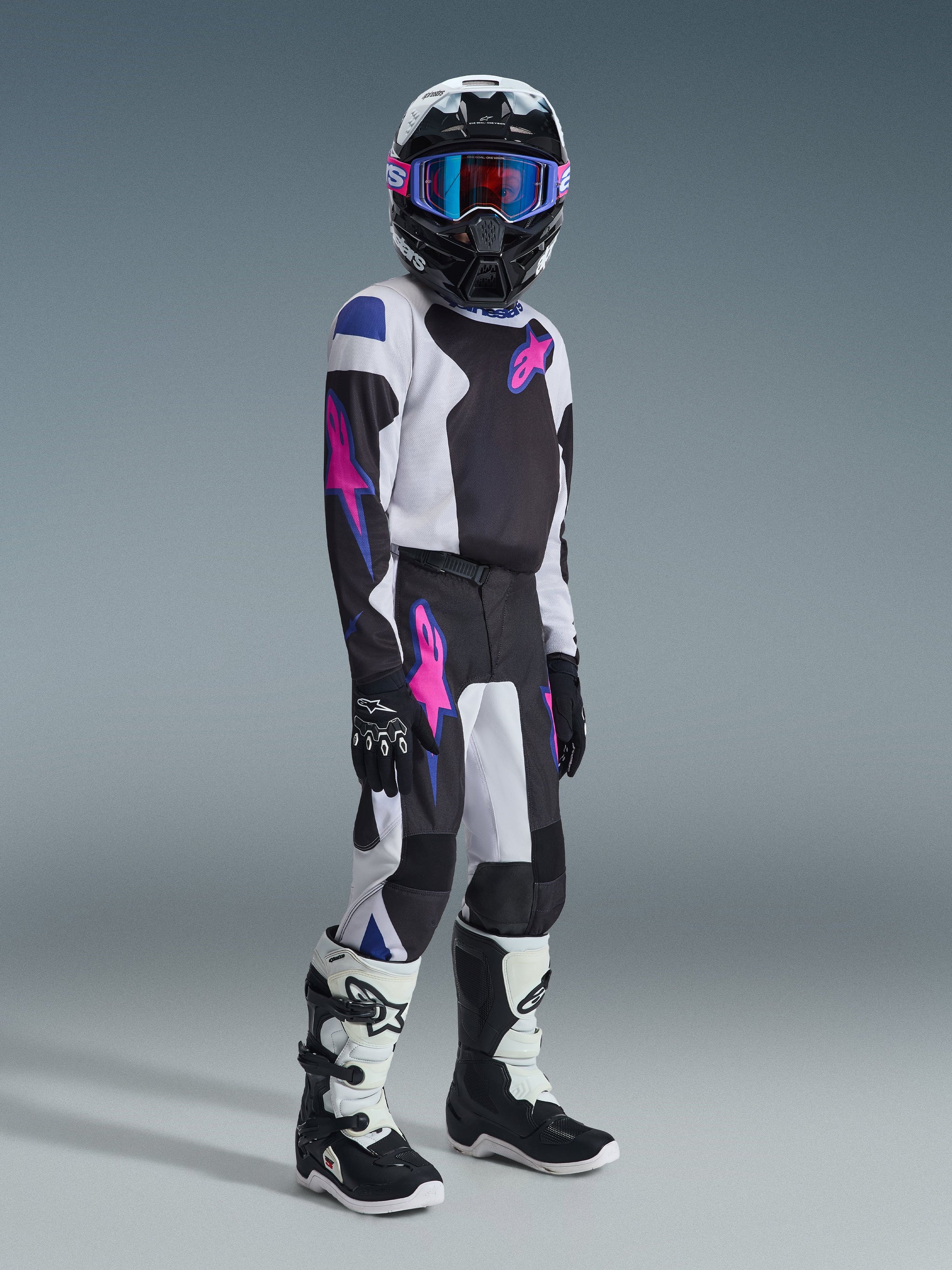 Young motocross rider wearing the Alpinestars 2026 Youth Fluid Grid Jersey in black, light gray, and purple, featuring pink star logos on the chest and sleeves. The outfit is paired with matching motocross pants, a black helmet with blue-tinted goggles, and black and white boots, representing a complete off-road racing kit for youth.